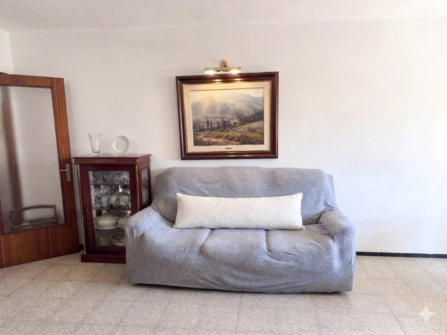 Living room with a gray sofa, white pillow, and framed landscape painting above. A glass cabinet stands to the left.