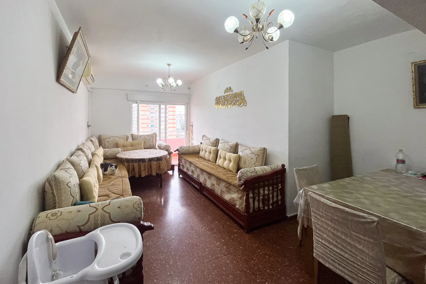 A bright living room with sofas, a table, and a high chair. A cat is resting on the sofa.