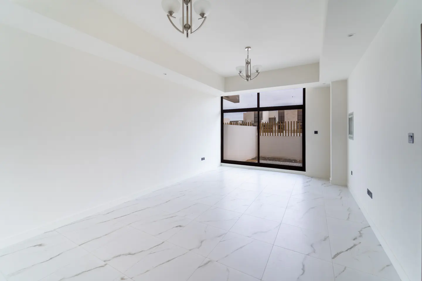 Bright, empty room with white walls, marble tile floor, and a large sliding glass door to a patio. Two chandeliers hang from the ceiling.