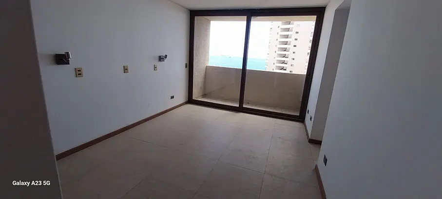 Empty room with beige tile floor, white walls, and brown trim. Sliding glass doors lead to a balcony with ocean and building views.