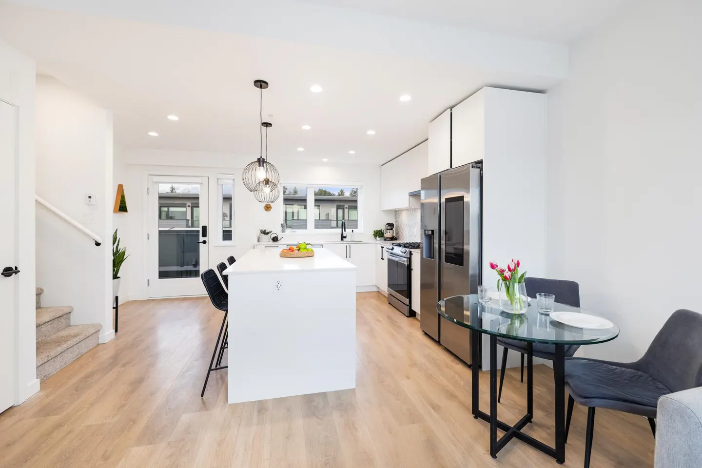 Bright, modern kitchen with white cabinets, stainless steel appliances, island with seating, and a glass dining table with gray chairs.