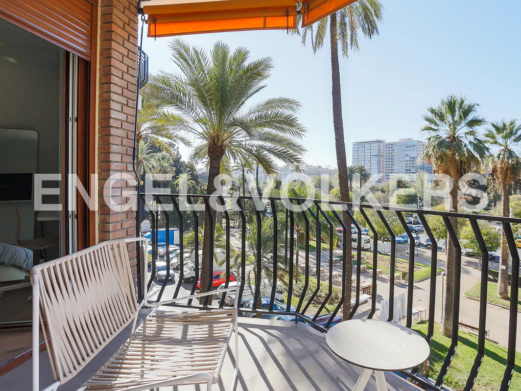 A balcony with a white woven chair and small round table, overlooking palm trees, a street, and city buildings.
