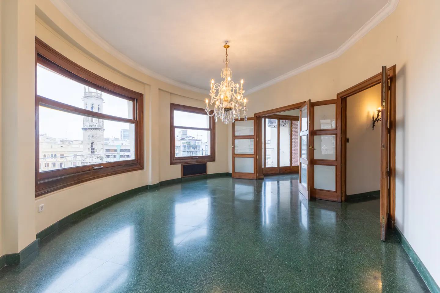 Empty room with green terrazzo floors, a crystal chandelier, and large windows overlooking a city with a clock tower.