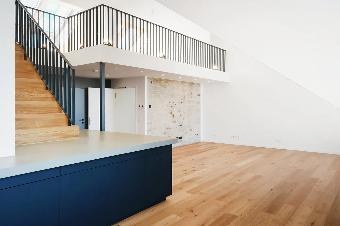 Bright, modern loft with wood floors, white walls, and a blue kitchen island. Stairs lead to a second-floor balcony with black railings.