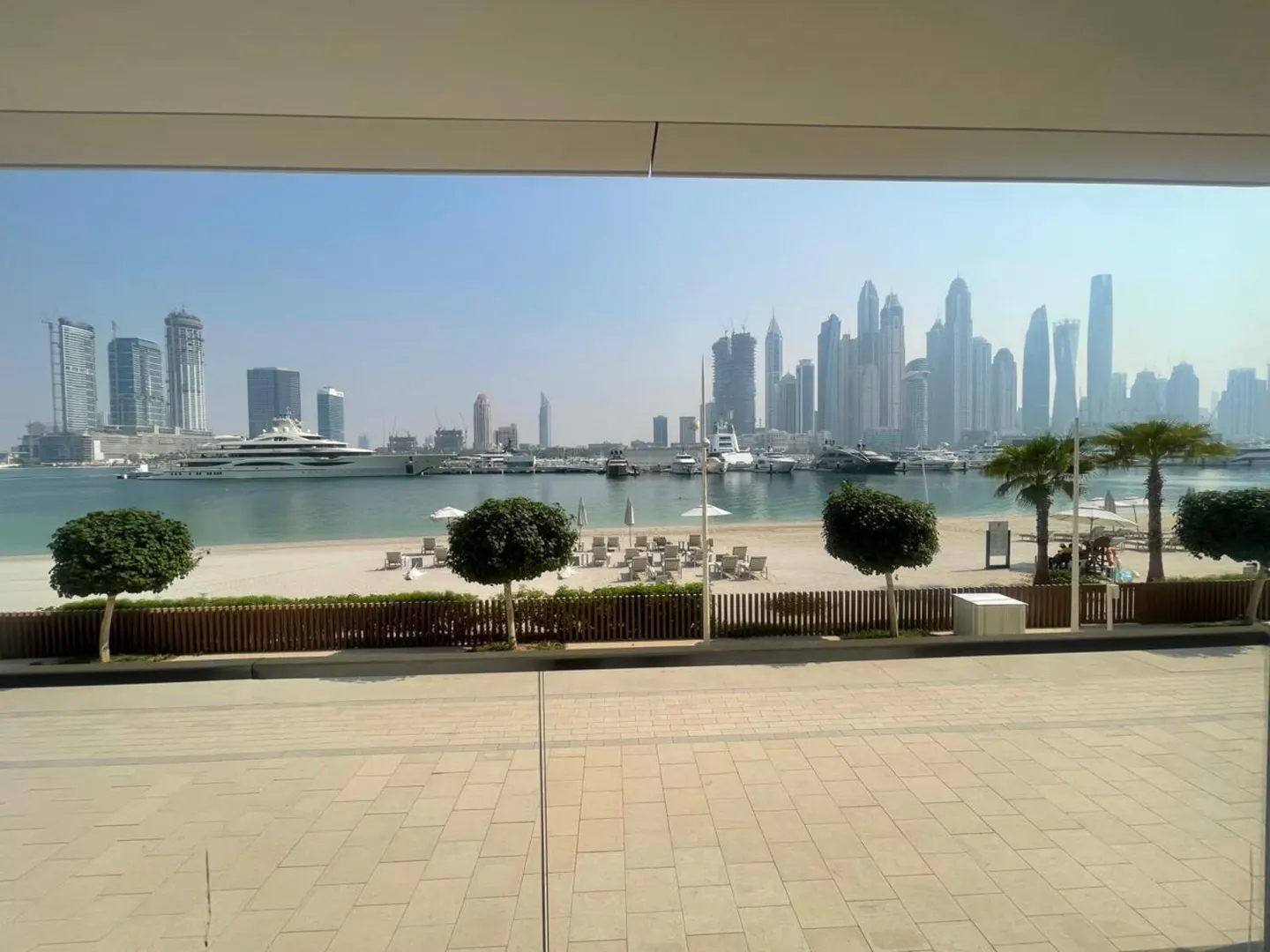 View from a balcony overlooking a sandy beach, palm trees, a calm harbor with yachts, and the Dubai skyline in the distance.
