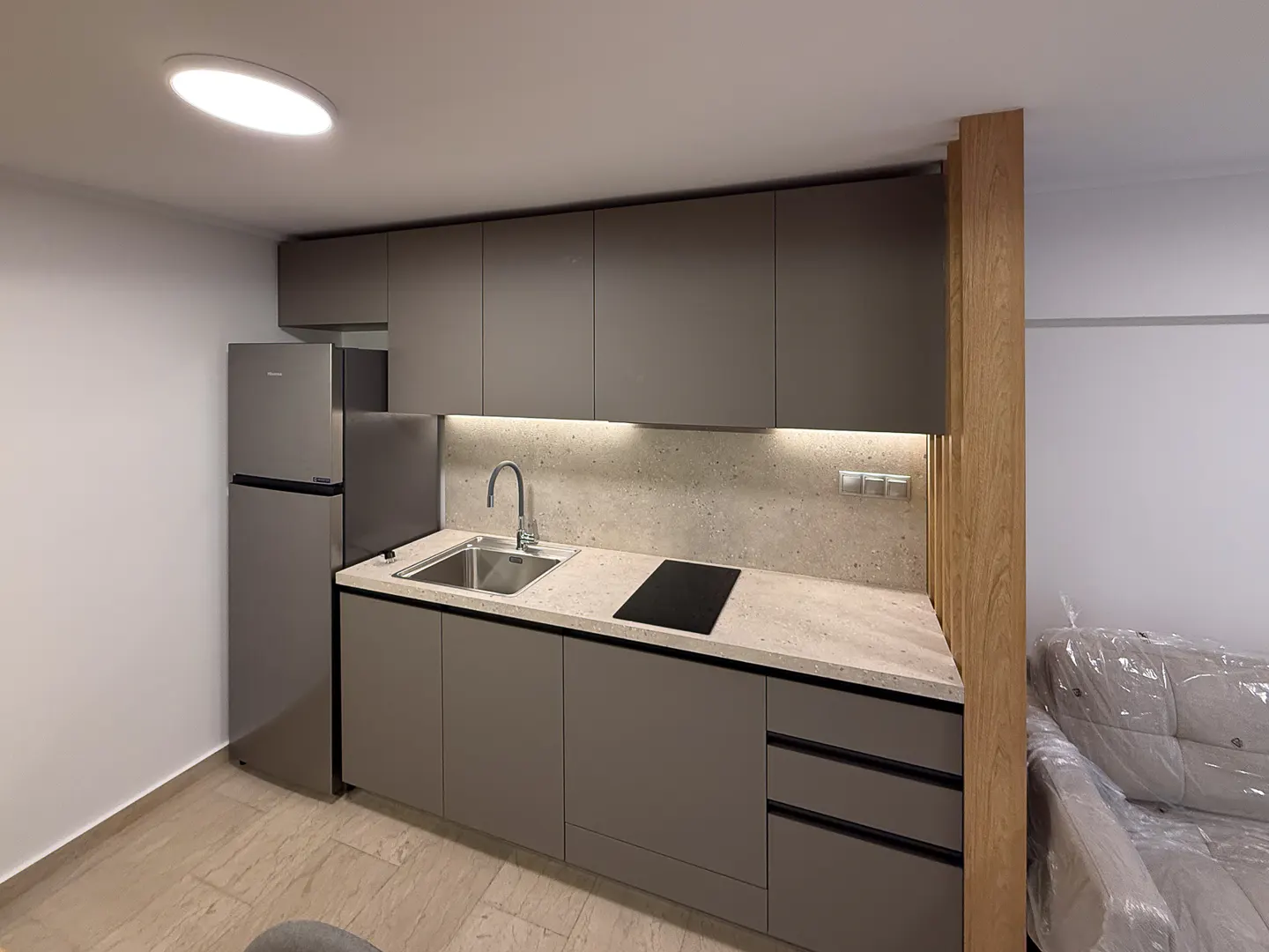 A modern kitchen with gray cabinets, stainless steel fridge, sink, and black cooktop. A light wood divider separates the kitchen from a sofa covered in plastic.