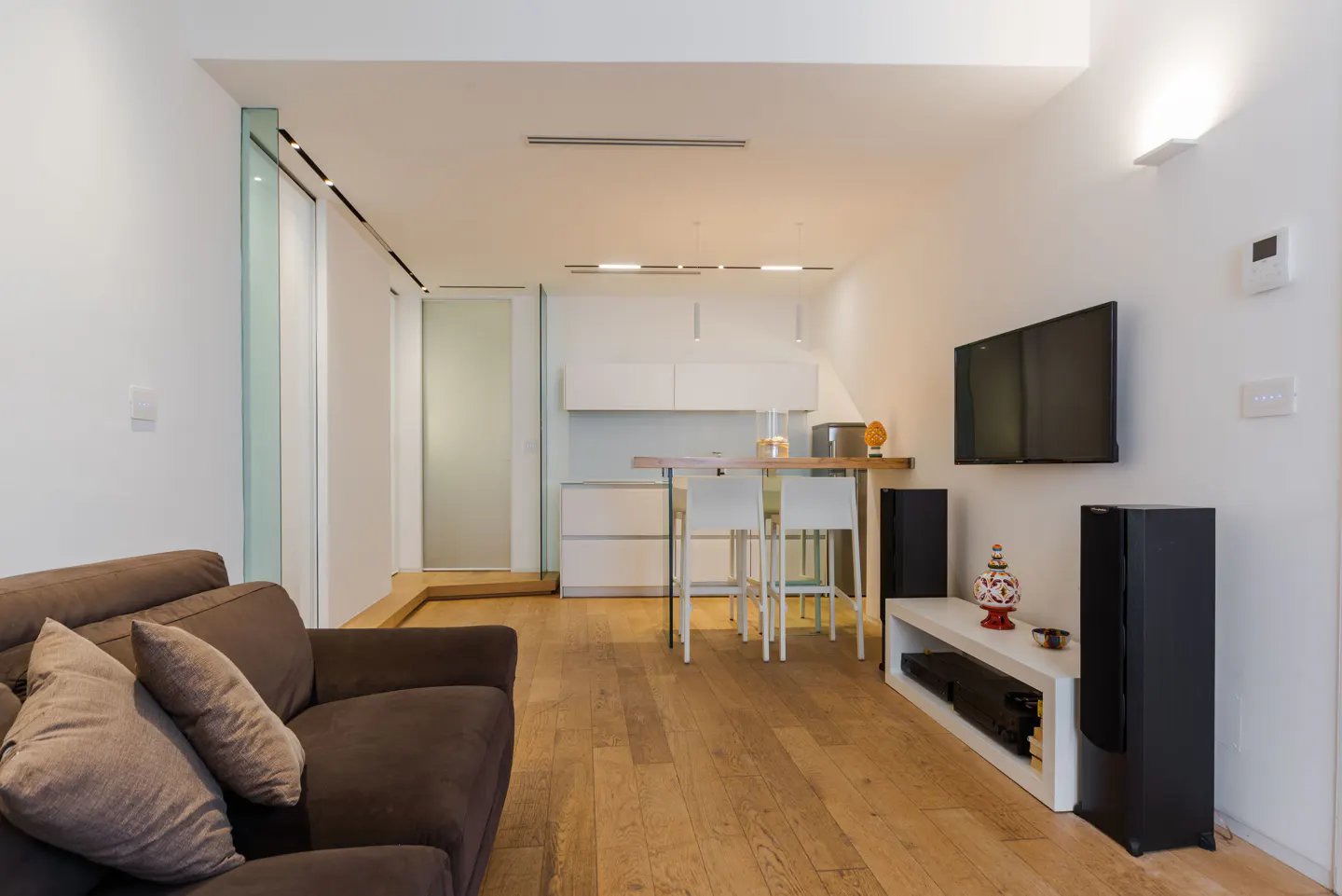 A modern living room with a brown sofa, TV, speakers, and a small kitchen area with white cabinets. The floor is light wood.