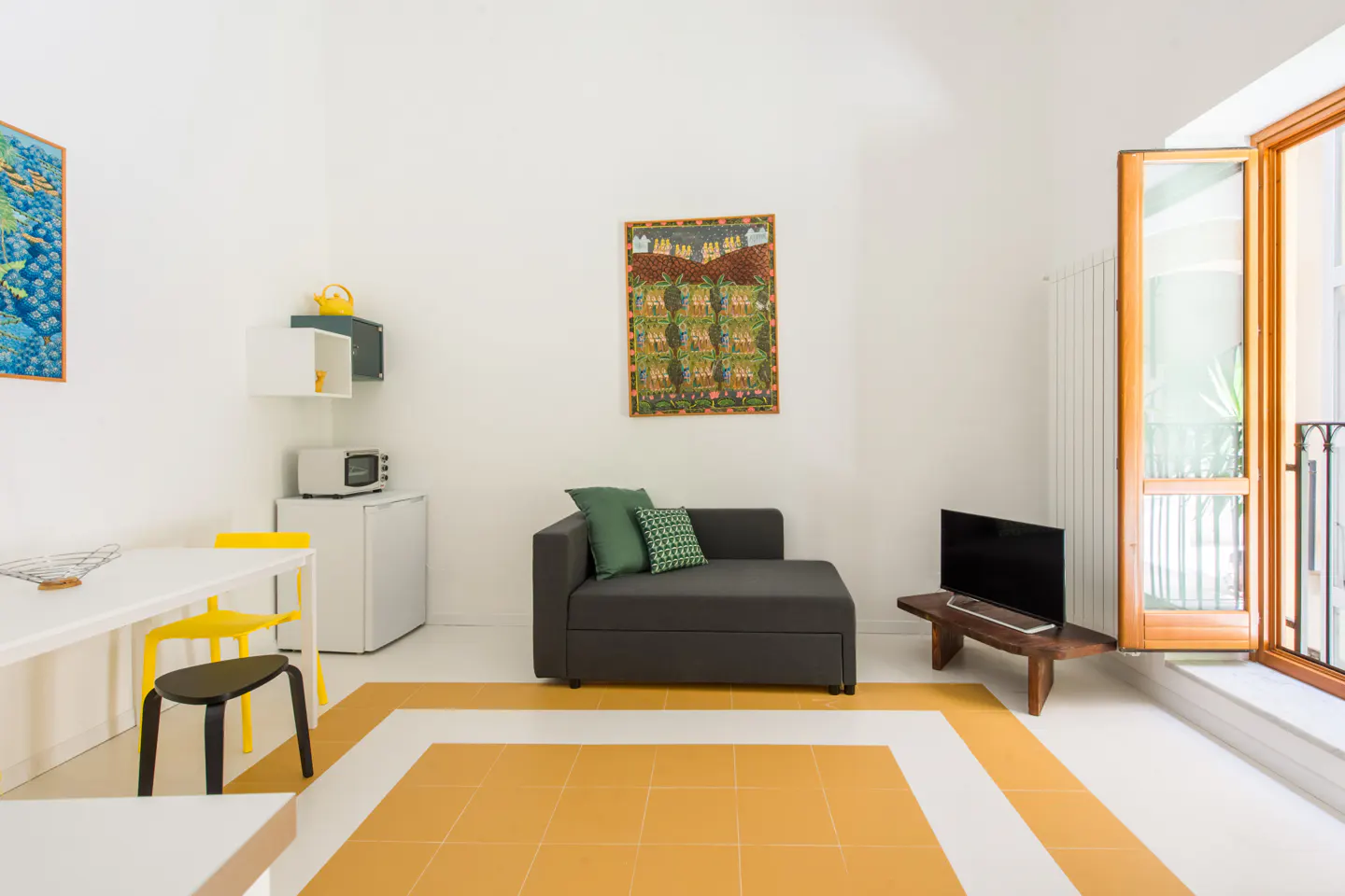 Bright, modern apartment interior with white walls, yellow tile border, gray sofa, and open balcony door. Artwork adorns the walls.