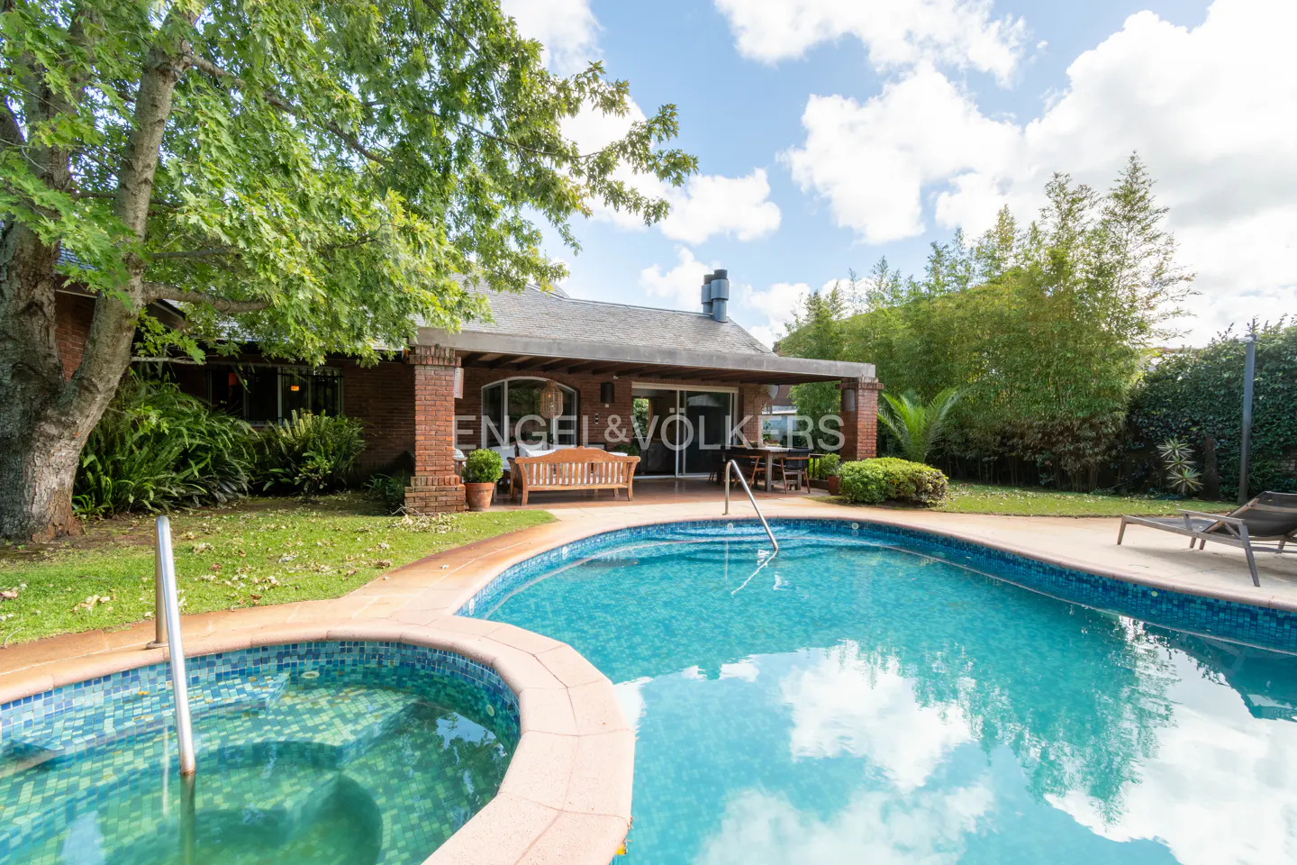 Backyard view of a brick house with a pool and jacuzzi on a sunny day. The Engel & Volkers logo is visible on the house.