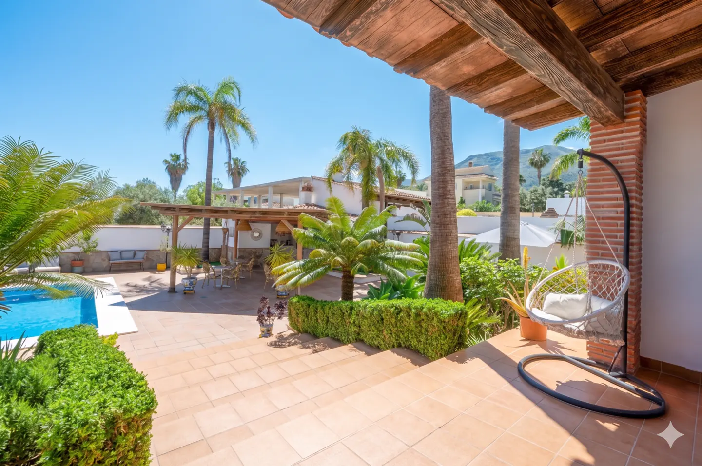 Outdoor patio with a hanging chair, pool, palm trees, and a wooden pergola under a clear blue sky.