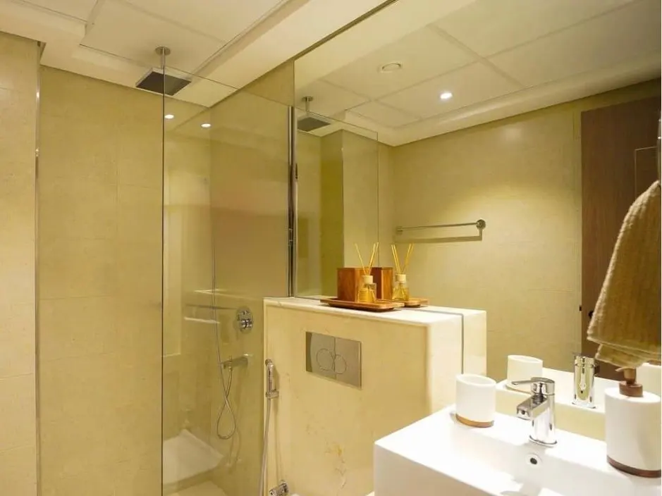 Bathroom with glass shower, white sink, and beige walls. Reed diffusers sit on a marble shelf. A towel hangs on the door.