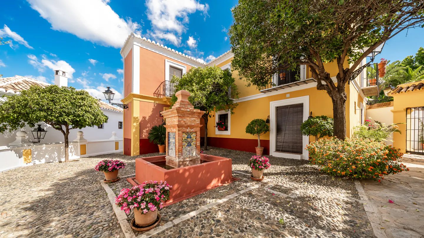 Cobblestone square with a red fountain, potted pink flowers, and yellow and peach buildings under a blue sky.