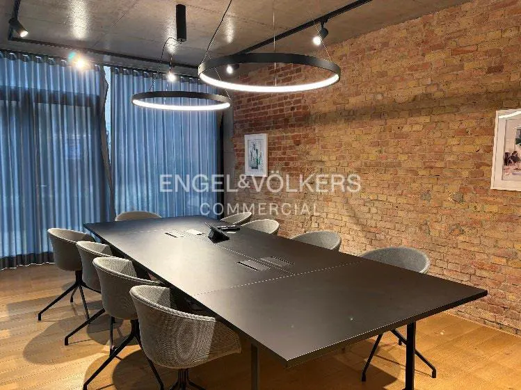 Conference room with a long black table, gray chairs, and circular lights. A brick wall and blue curtains are in the background.