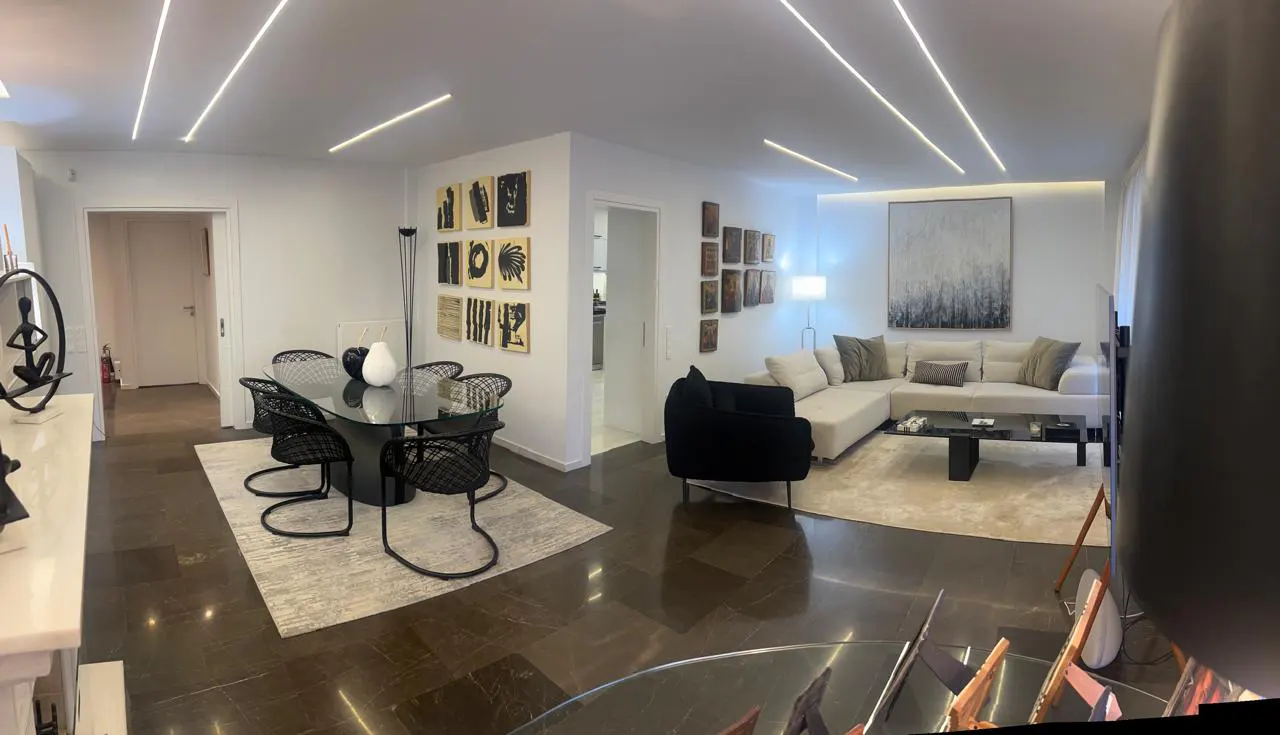 A modern apartment interior with a dining area featuring a glass table and black chairs, a living room with a beige sectional sofa, and dark marble floors.