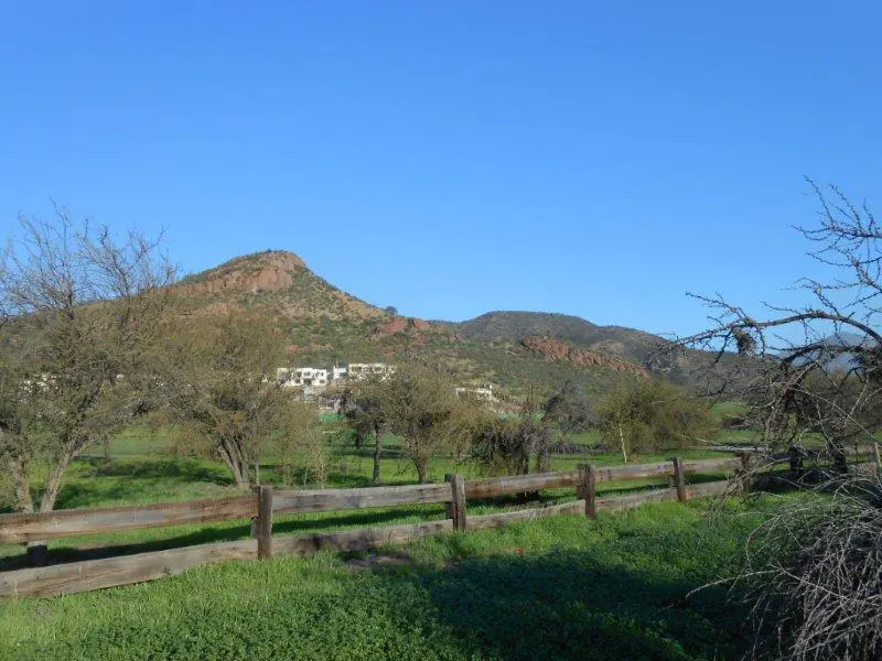 Landscape view of a green field with a wooden fence, mountains, and a clear blue sky.