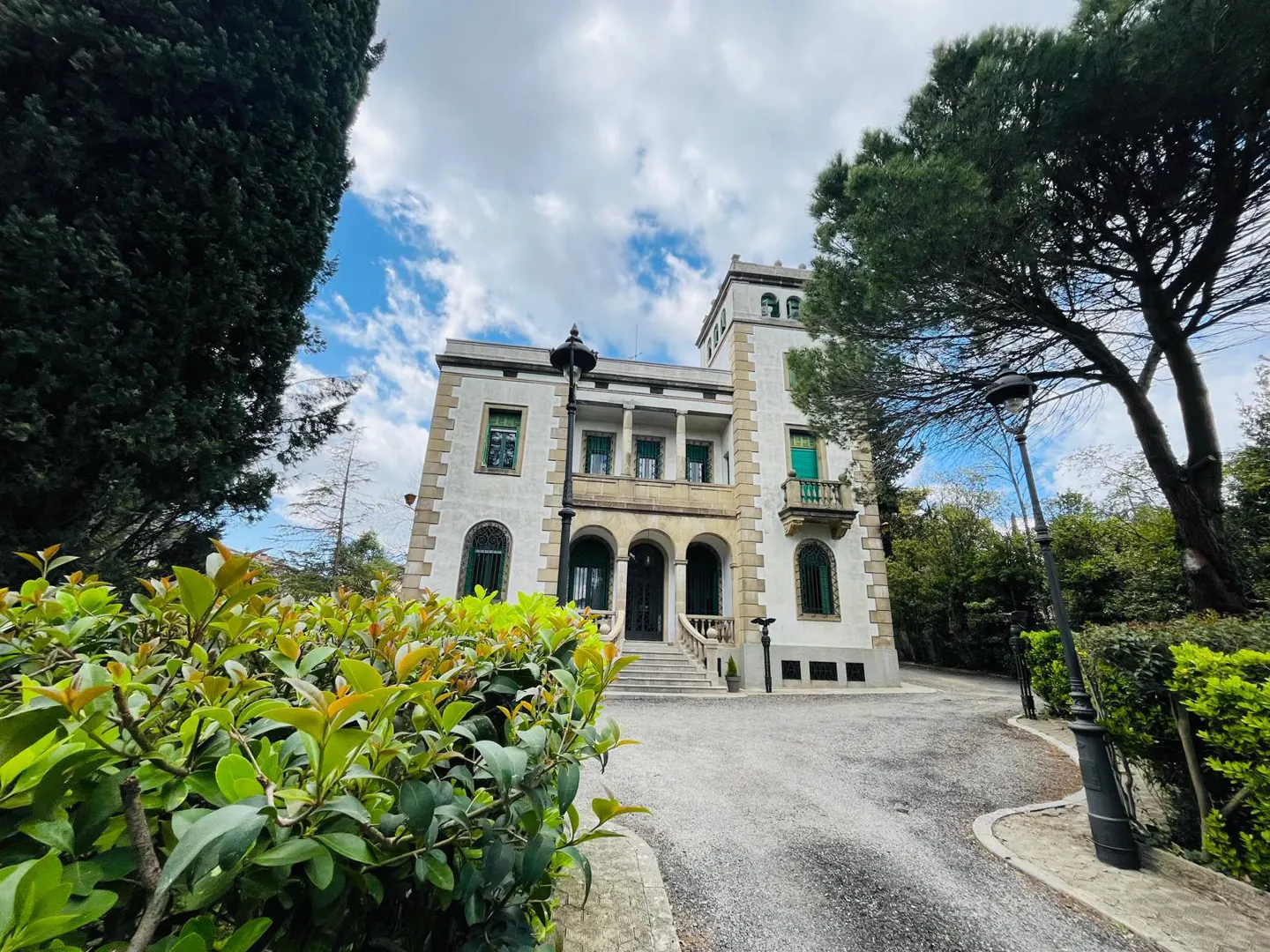 Exterior of a large, white, multi-story house with green trim, a stone facade, and a tower, surrounded by trees and greenery.