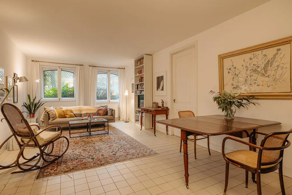 Bright living room with a rocking chair, sofa, rug, and dining table with chairs. A painting hangs on the wall.