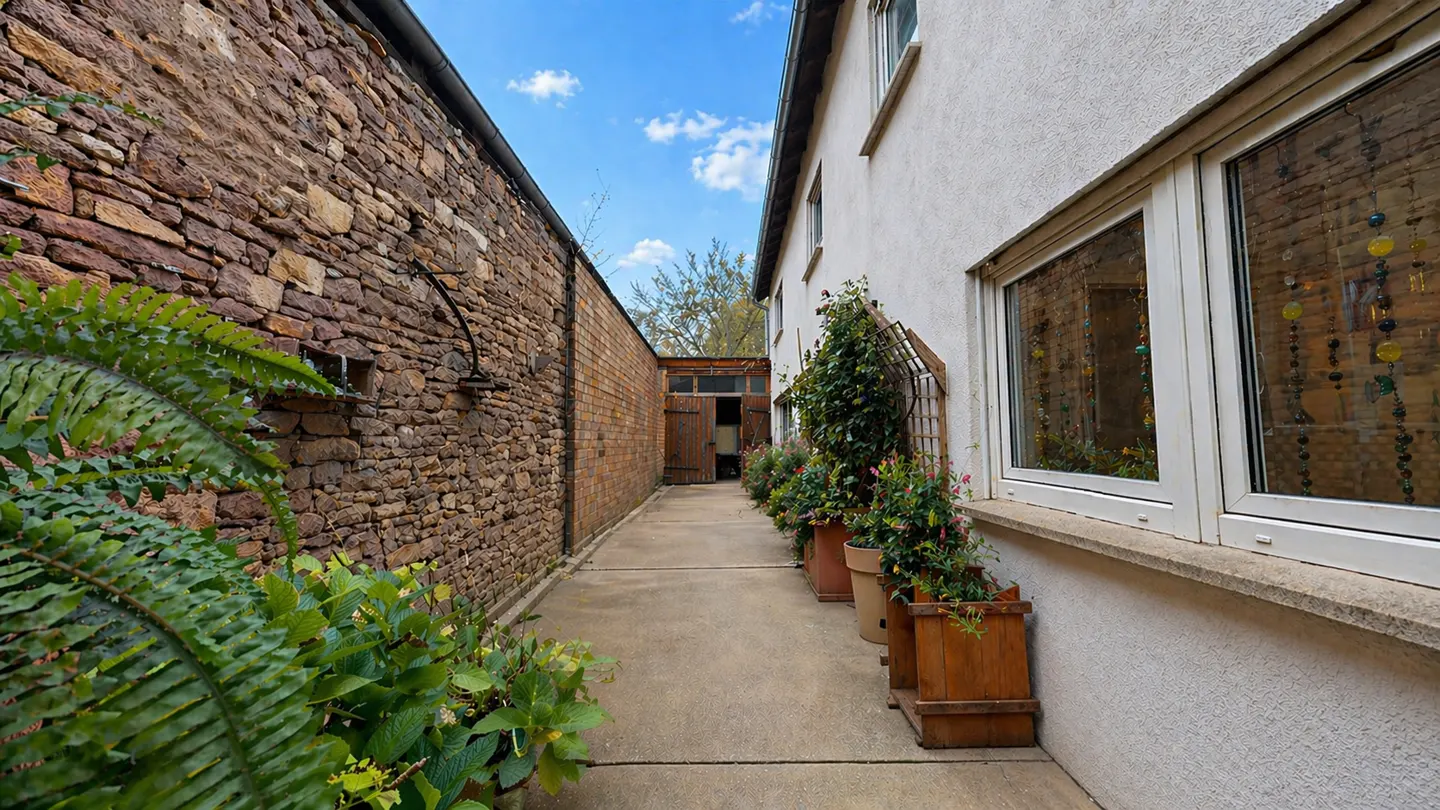 Narrow walkway between a stone wall and a white building with potted plants.