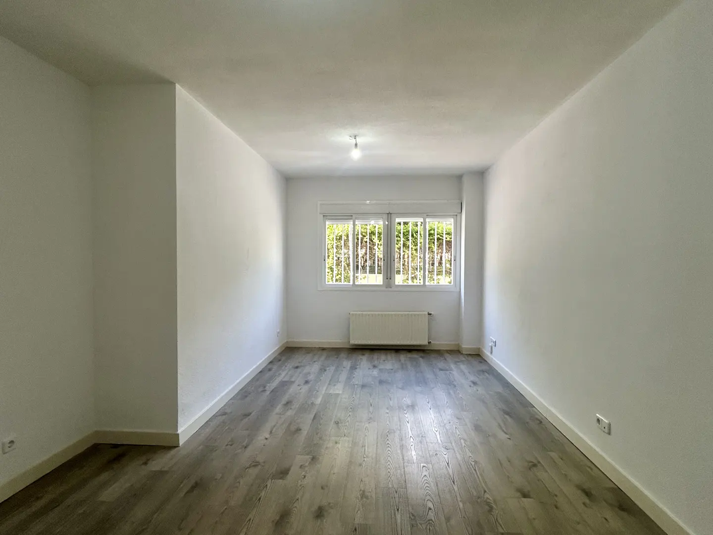 Empty room with white walls, gray wood floor, and a window with bars overlooking green trees.