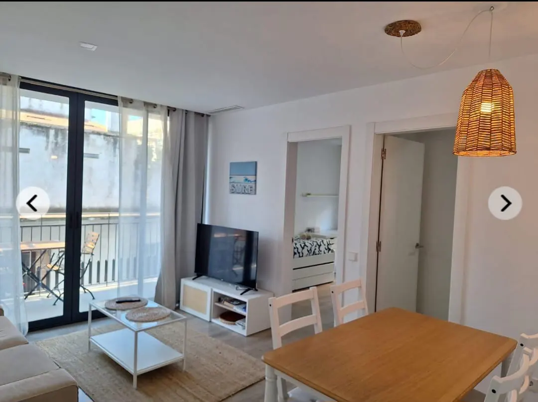 Bright living room with a balcony, a TV, a dining table, and a woven pendant light. The walls are white, and the furniture is light-colored.