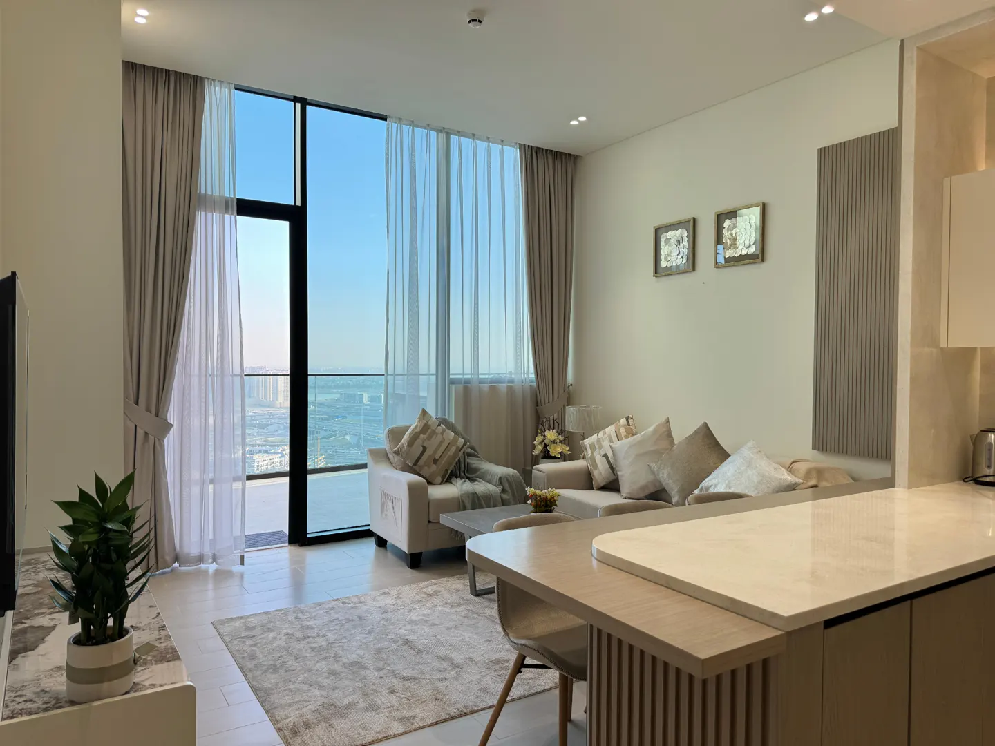 Living room with beige sofa, rug, and floor-to-ceiling windows with city views. Kitchen island in the foreground.