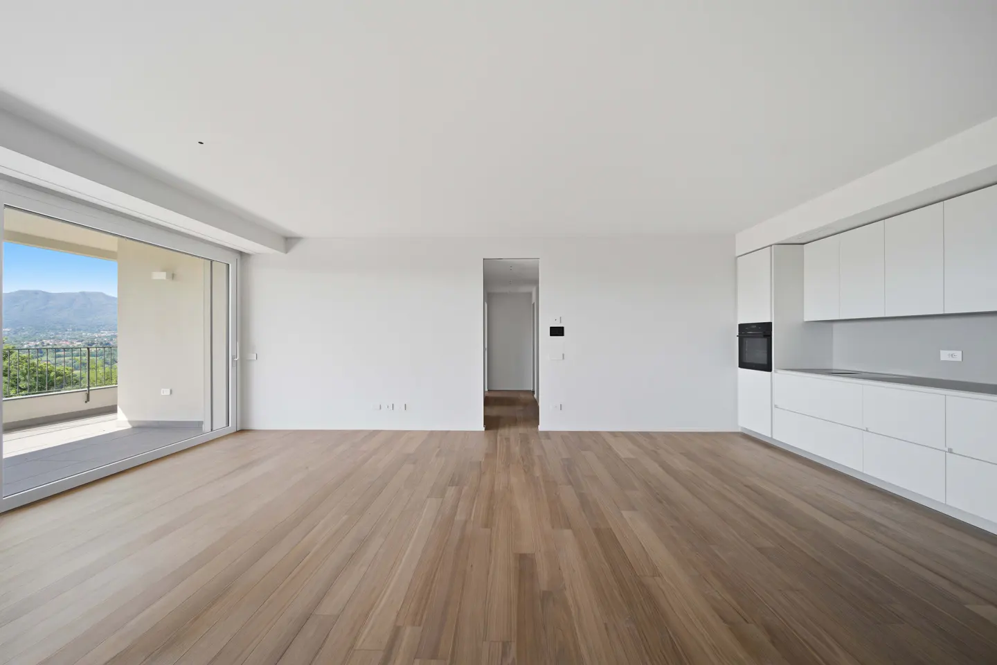 Bright, empty room with wood floors, white walls, and white kitchen cabinets. A sliding glass door leads to a balcony with a mountain view.