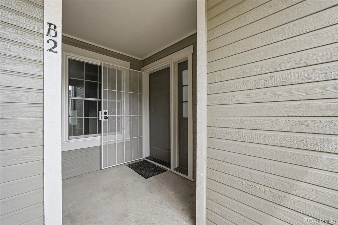Exterior view of unit B2 with a gray door, white security screen, and beige horizontal siding.