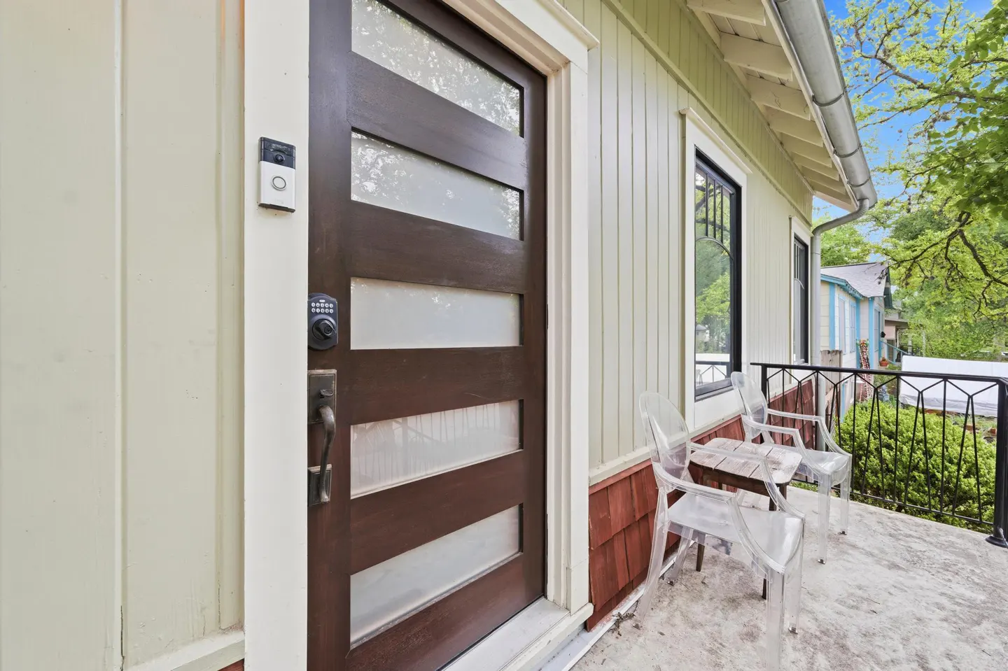 Exterior view of a house with a brown front door with glass panels, a Ring doorbell, and a small porch with clear chairs and a table.