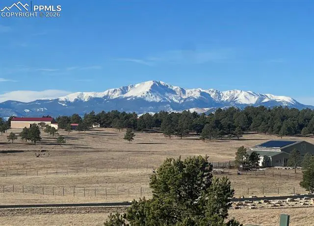 Scenic view of snow-capped mountains, green trees, and a brown field under a clear blue sky. Buildings are visible in the distance.