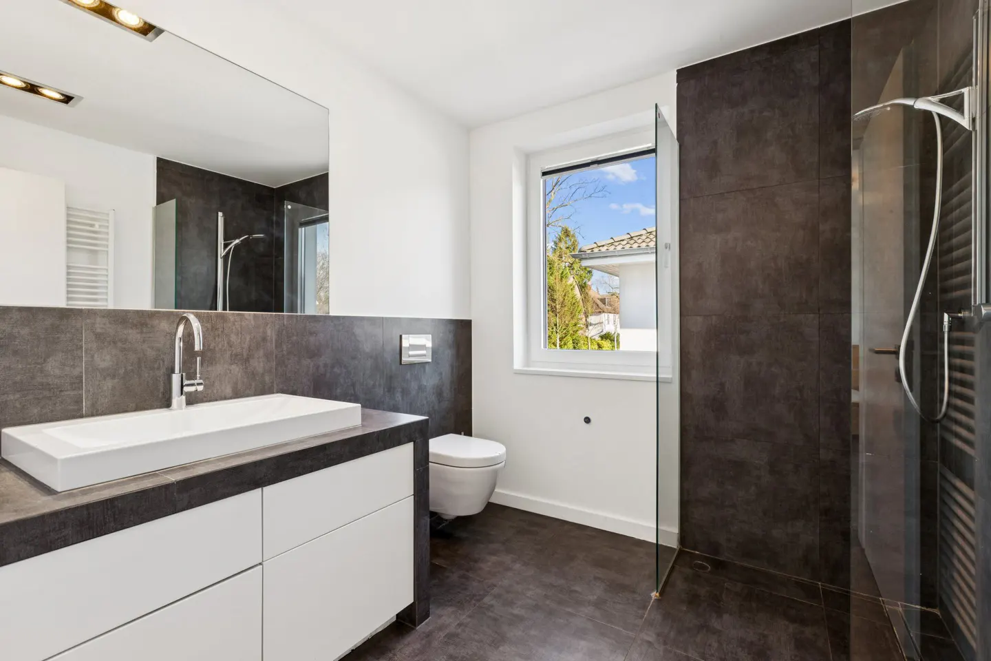 Modern bathroom with white walls, dark gray tiles, and a white vanity with a rectangular sink. A glass shower door and a window with a view are also visible.