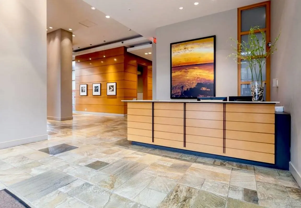 Hotel lobby with a light wood reception desk, stone tile floor, and a painting of a sunset on the wall.