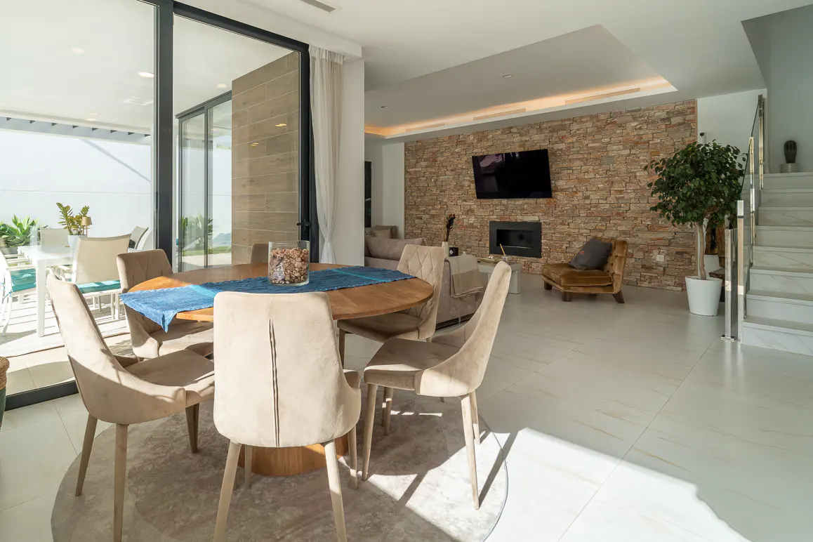 Bright, modern dining area with a round wooden table, beige chairs, and a blue runner. A stone accent wall features a TV and fireplace. Sliding glass doors lead to an outdoor patio.