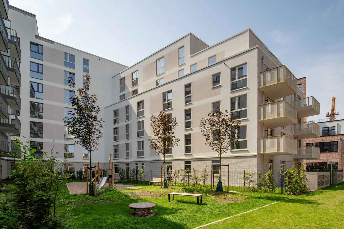Apartment complex with a playground. The building is white with balconies and many windows. The playground has a slide and benches.