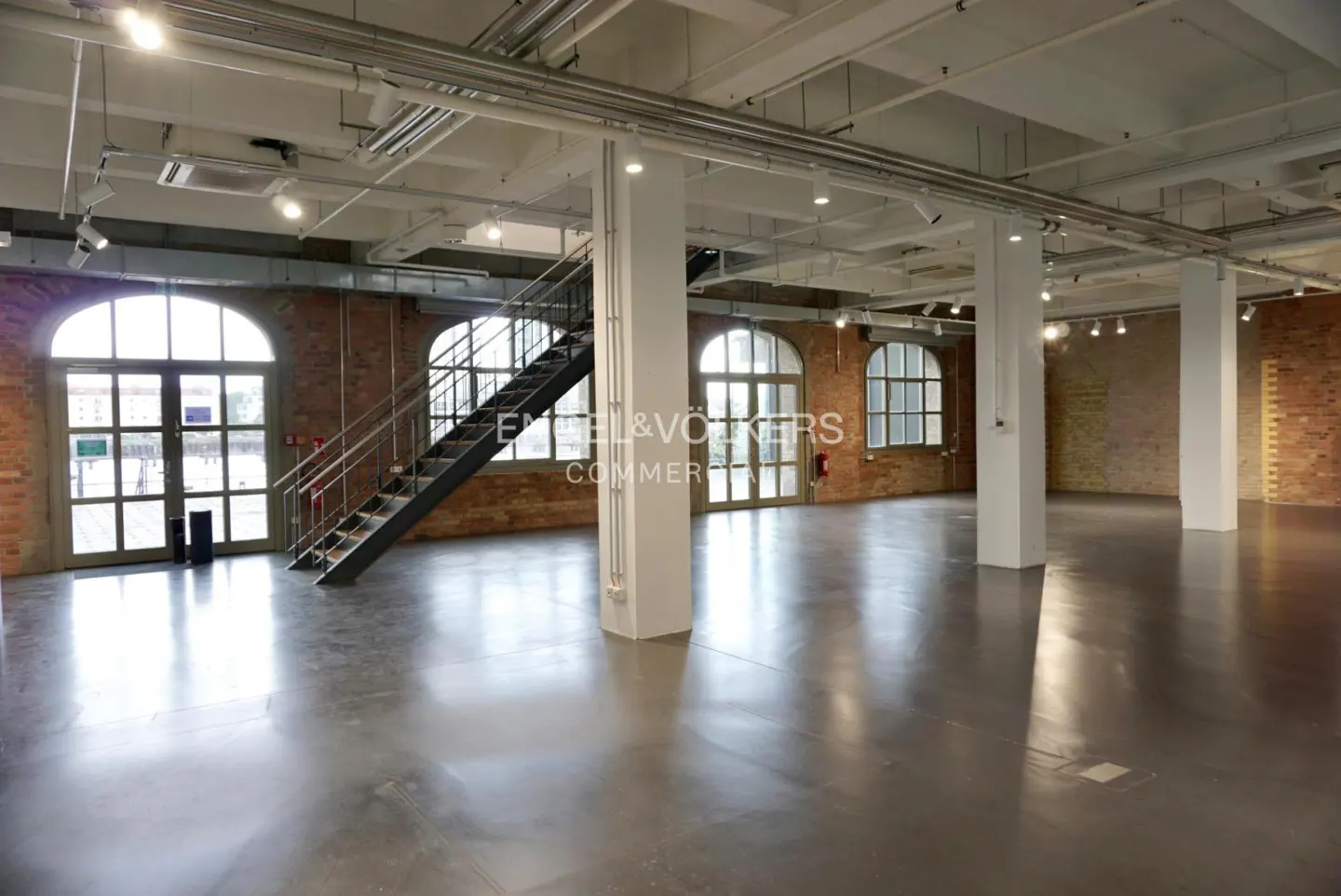Bright, empty commercial space with polished concrete floors, brick walls, and large arched windows. White support columns and exposed ceiling pipes.