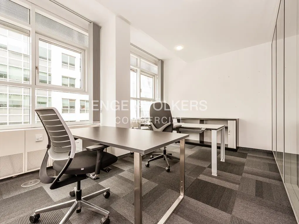 Bright office space with gray desks, black chairs, and large windows. The floor is carpeted in a gray geometric pattern.