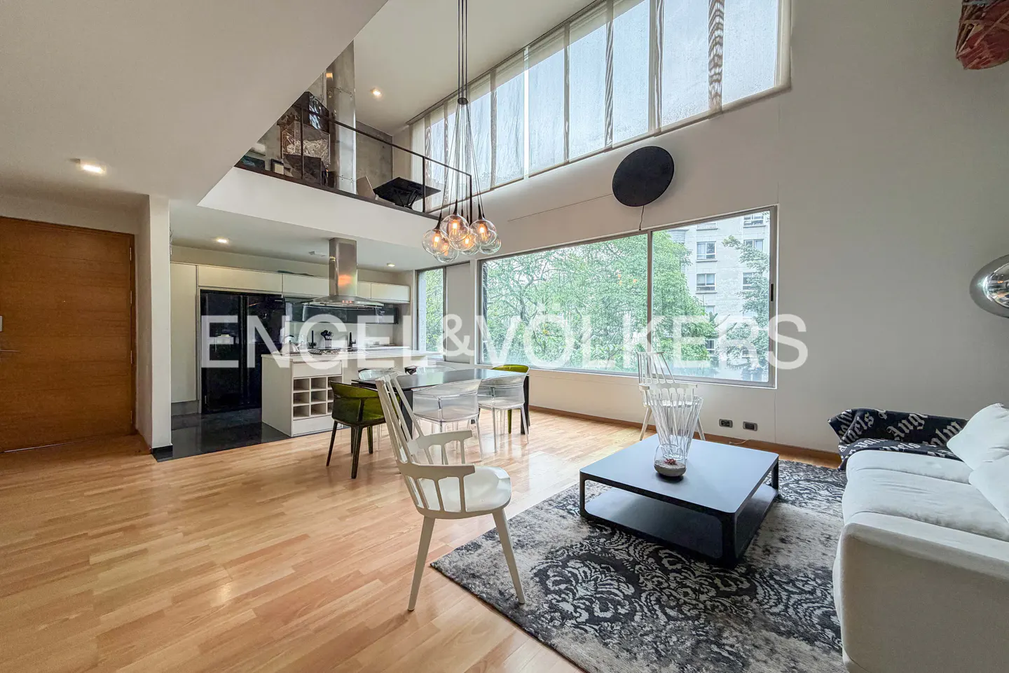 Bright, modern apartment interior with wood floors, white sofa, and large windows overlooking greenery. Kitchen and dining area visible.