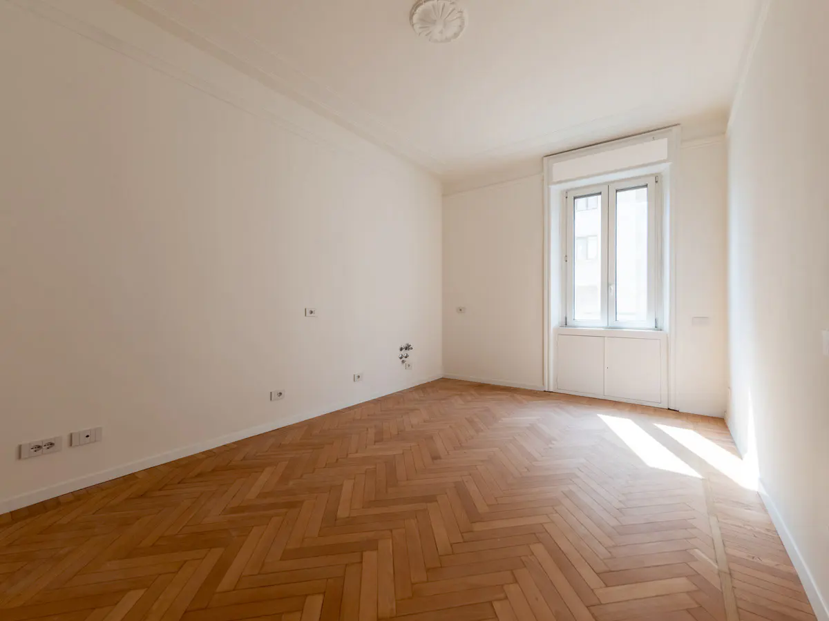 Empty room with white walls, herringbone wood floors, and a window letting in natural light.
