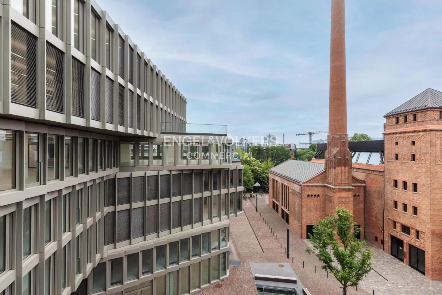 Exterior view of a modern, gray building next to a brick building with a tall chimney. "Engel & Völkers Commercial" is visible on the modern building.