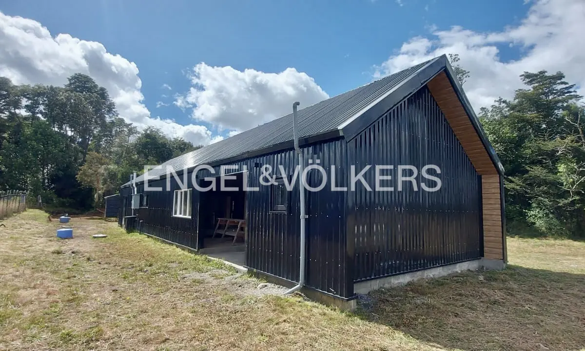Black corrugated metal barn-style house with a gray roof, surrounded by green grass and trees under a blue sky.