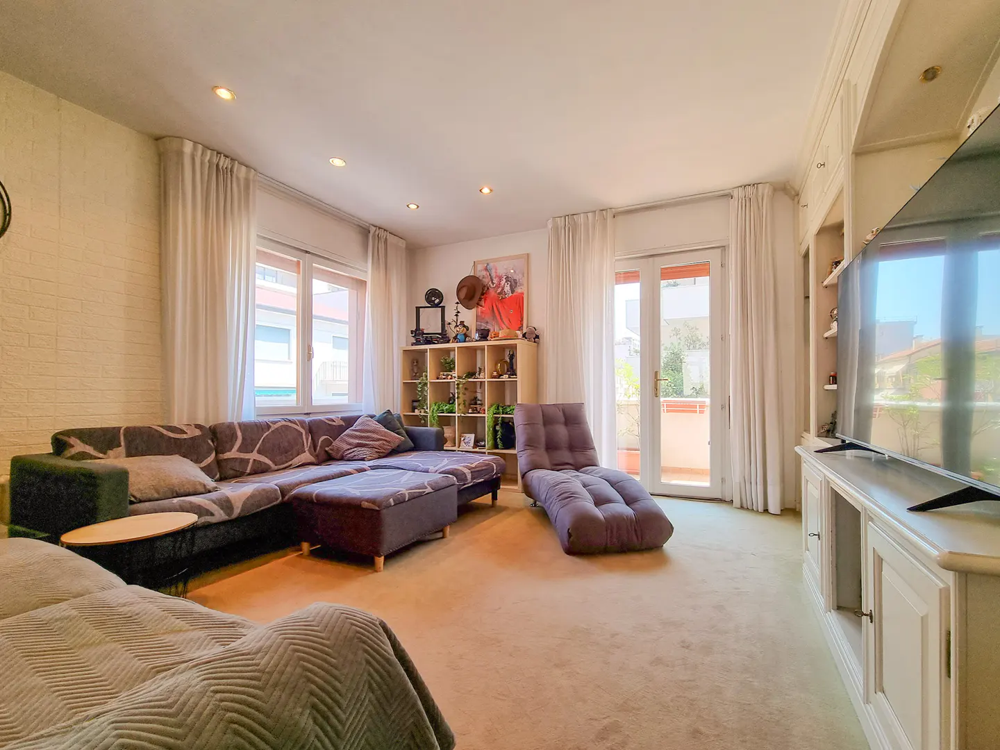 Bright living room with a large grey sectional sofa, a floor lounge chair, a wooden shelving unit, and a large TV.