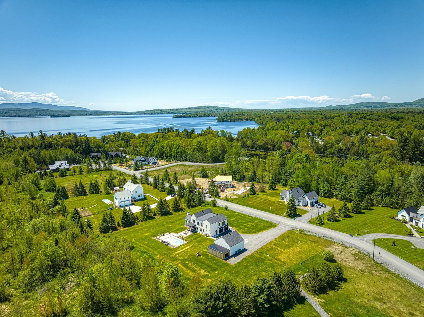 Aerial view of homes with green lawns near a lake and forest under a blue sky.