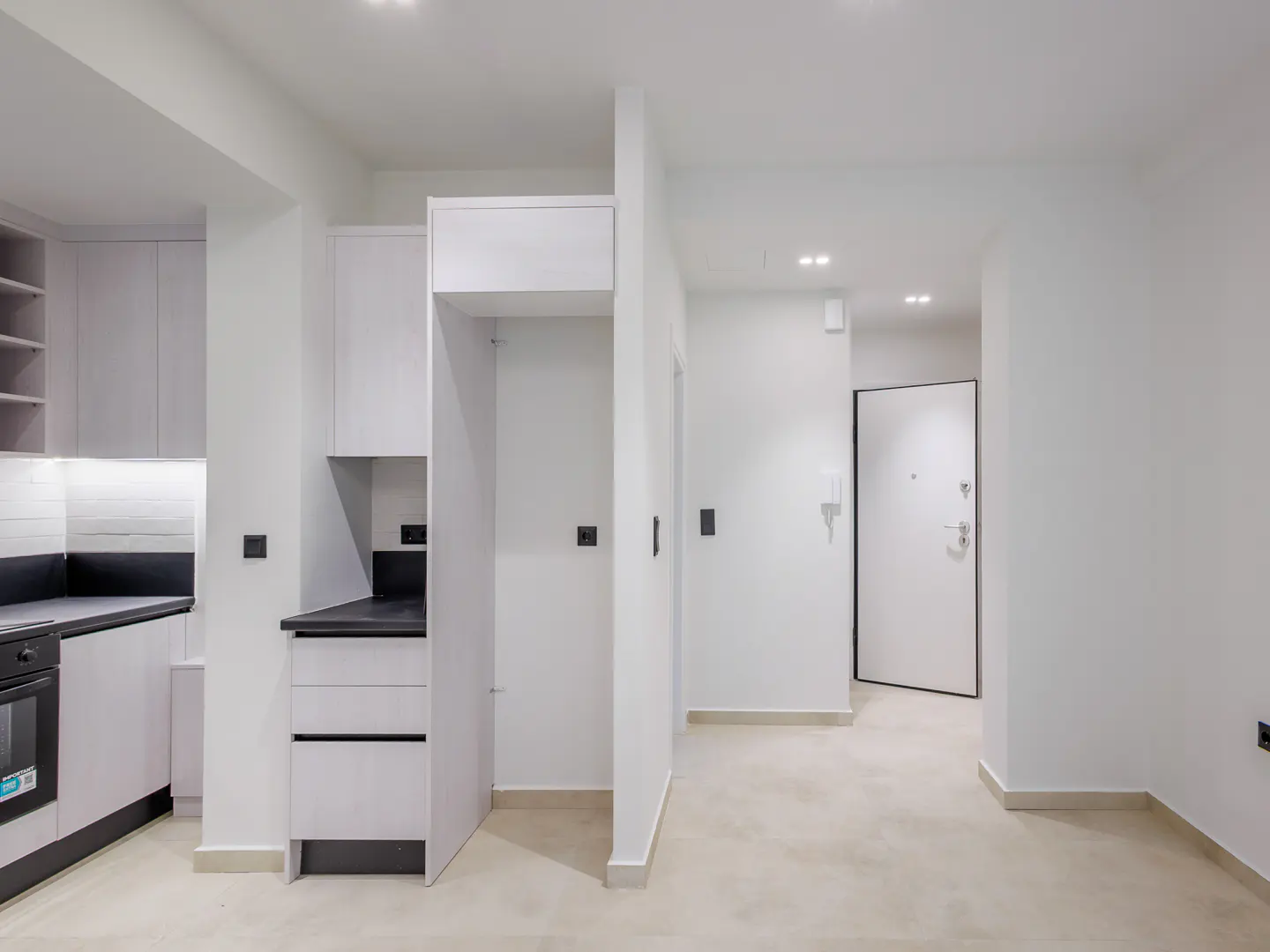 Bright, modern apartment interior with white walls, cabinets, and a black countertop. An open doorway leads to a hallway with a white door.