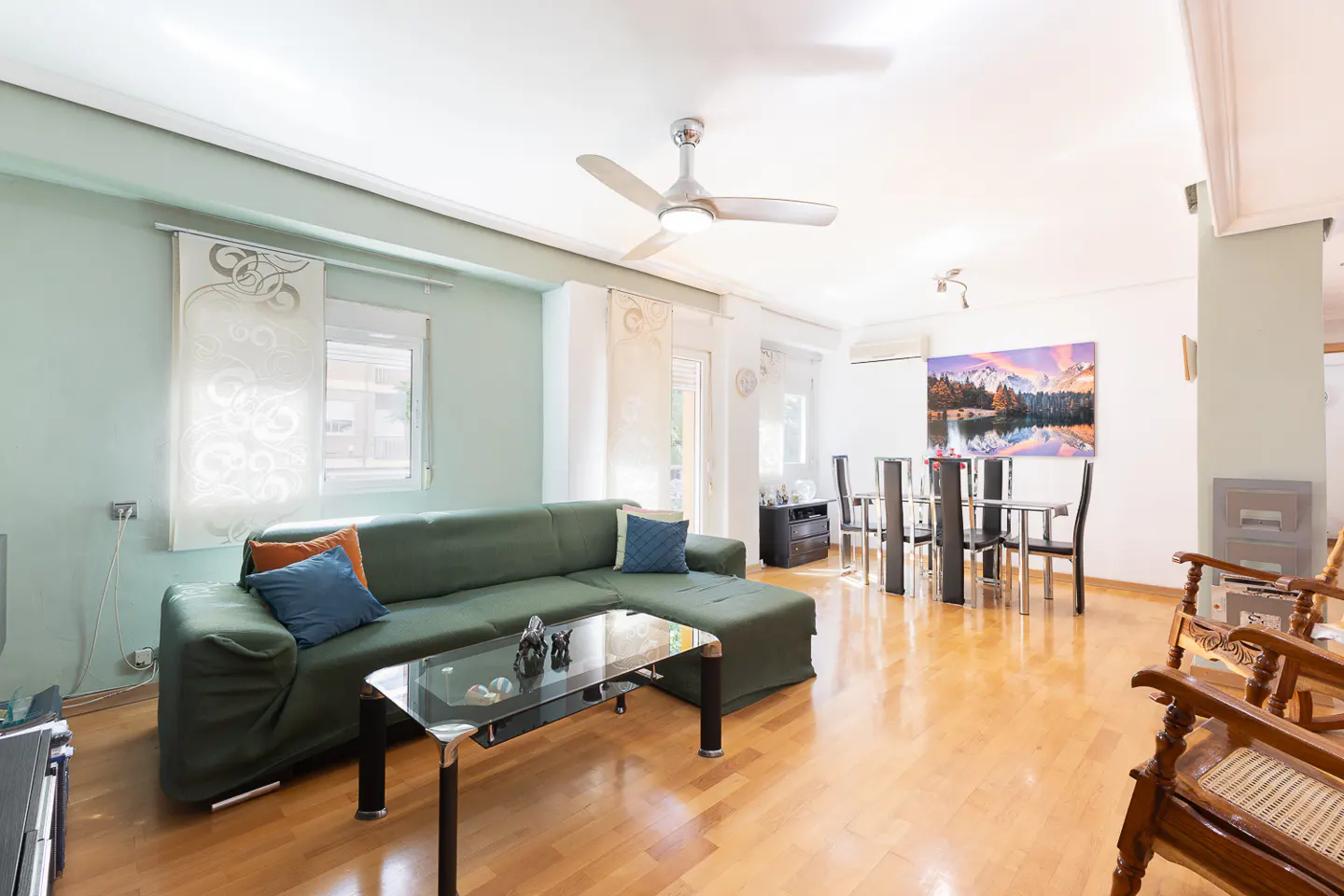 Bright living room with a green sectional sofa, glass coffee table, and a dining area with a mountain landscape painting.