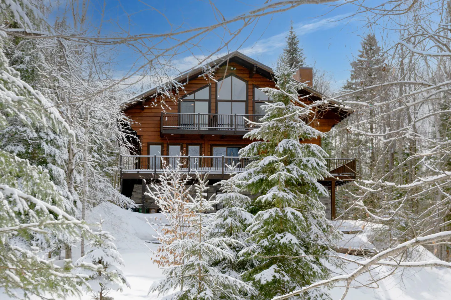 A two-story log cabin with balconies is surrounded by snow-covered trees under a blue sky.