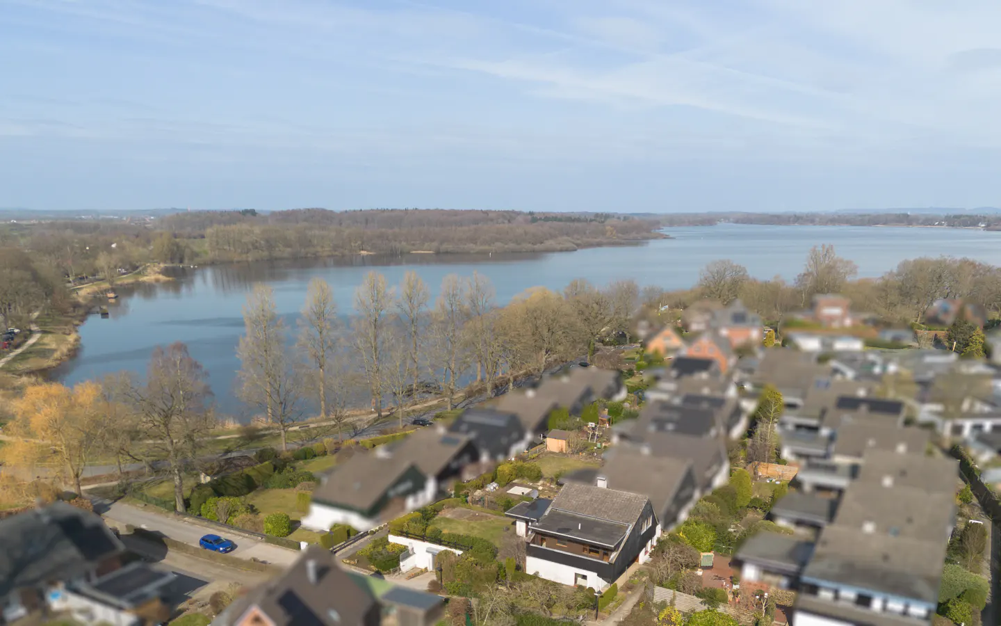 Aerial view of houses near a lake under a blue sky, with trees lining the shore.