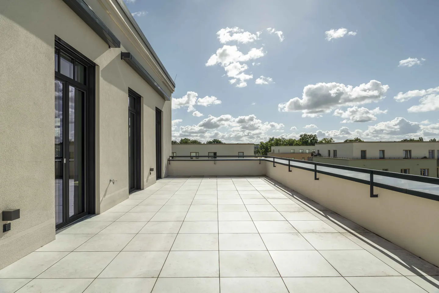 A rooftop patio with light gray tiles, black railings, and black-framed windows under a partly cloudy sky.