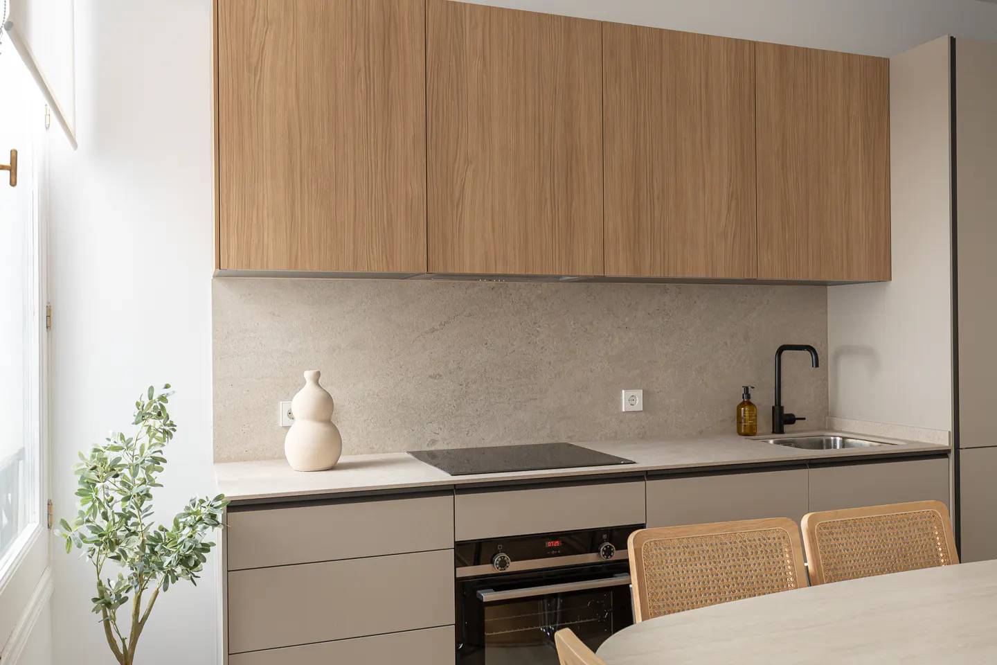 Modern kitchen with light wood cabinets, beige countertops and backsplash, black faucet, and woven chairs.