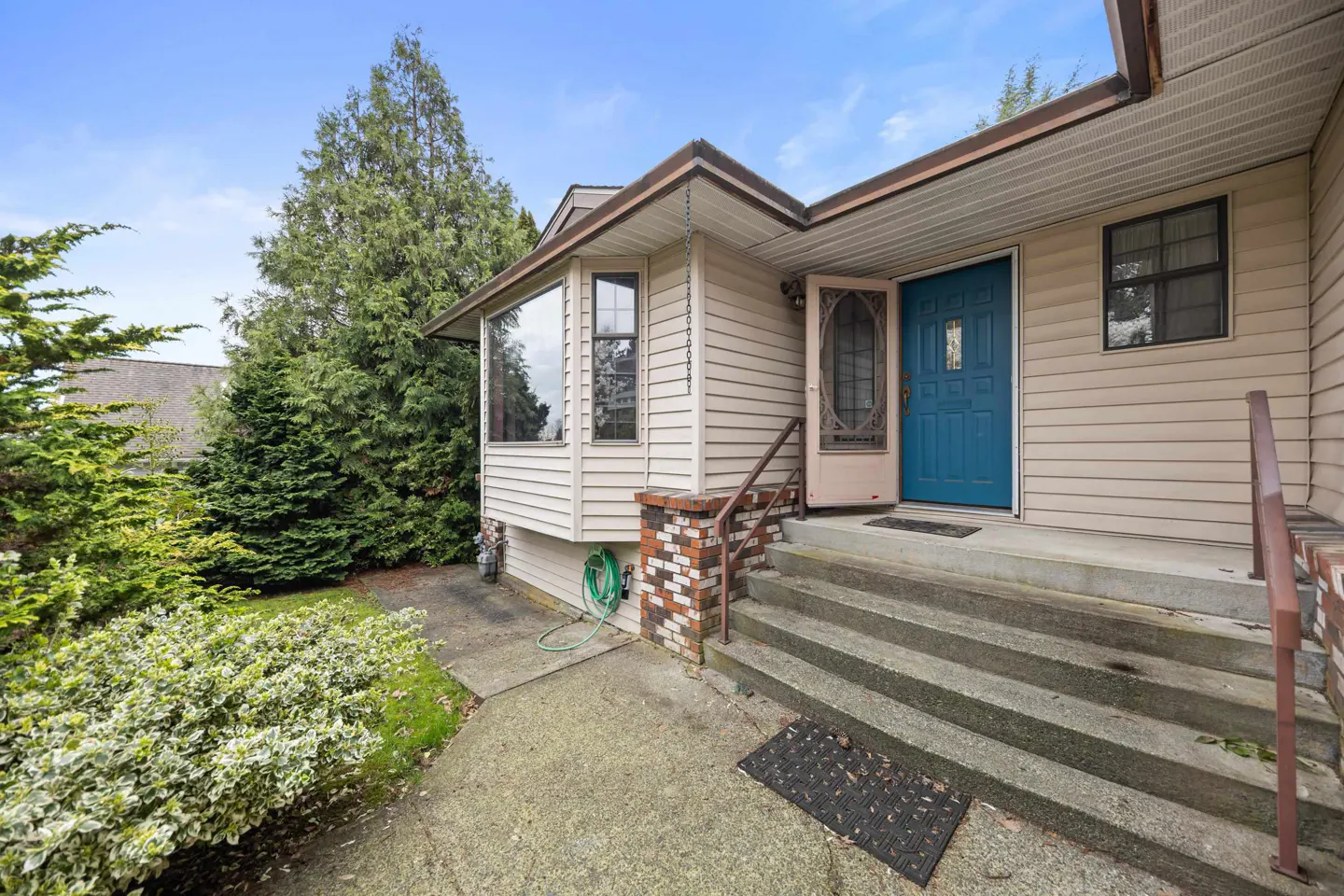 Exterior view of a home with beige siding, a bright blue front door, concrete steps, and a brick-accented porch.
