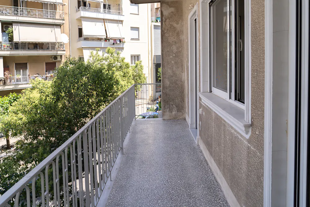 A long, narrow balcony with a gray metal railing overlooks trees and buildings. White-framed windows are on the right.