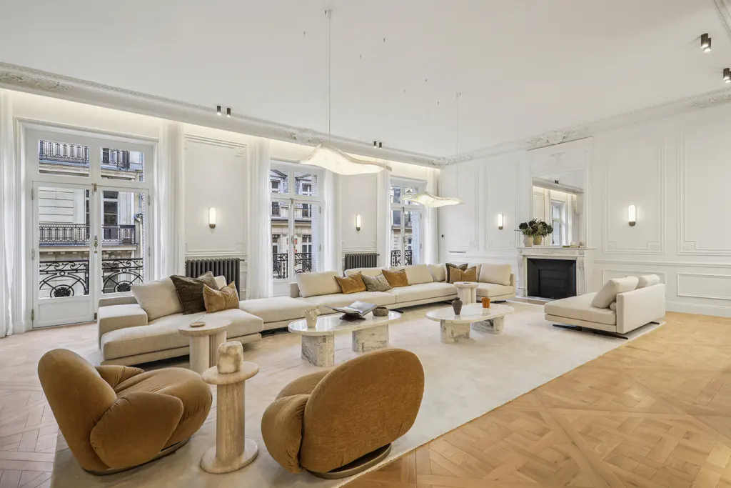 Bright, spacious living room with white walls, large windows, and light wood floors. Beige sectional sofa, brown armchairs, and marble tables.